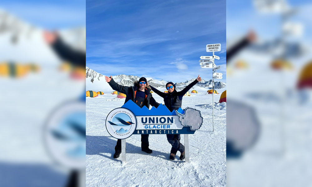 Chris and Julie, in snow covered Antarctica, pose behind a sign which reads: Union Glacier, Antarctica.