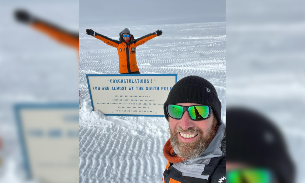 Chris and Julie standing and smiling beside a sign that reads: Congratulations! You are almost at the South Pole.